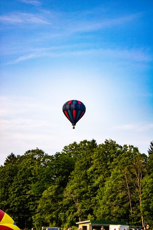A colorful hot air balloon soars above lush green trees against a bright blue sky.