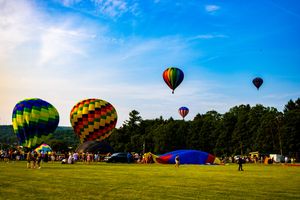 Colorful hot air balloons launch and float over a green field at a sunny balloon festival with crowds below.