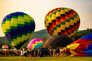 Colorful hot air balloons inflate on a green field at dusk during a balloon festival with mountains in the background.