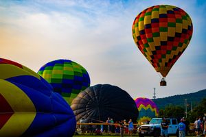 Colorful hot air balloons inflate and soar at a festival, with one multicolored balloon flying above a green field at dusk.