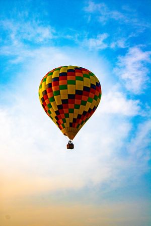 Colorful checkered hot air balloon soaring high in a bright blue sky with wispy white clouds.