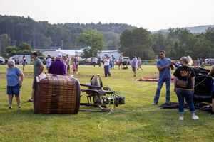 People gather around a large wicker hot air balloon basket on a green field at an outdoor festival event.