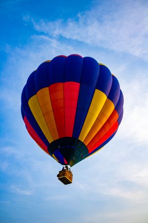 Colorful hot air balloon with blue, red, yellow stripes soaring against a bright blue sky with wispy clouds.