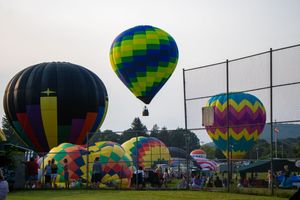 Colorful hot air balloons launching at a festival on a grassy field near a baseball fence at dusk.
