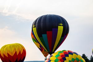 Colorful hot air balloons float against a cloudy sky at a balloon festival, with a large dark blue balloon in the center.