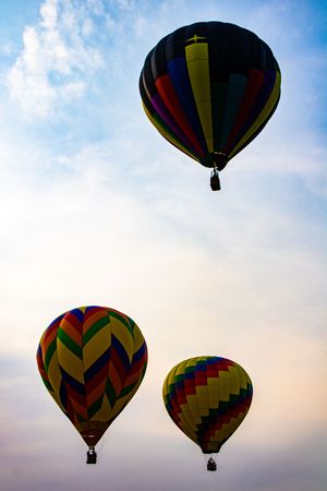 Three colorful hot air balloons with chevron patterns float against a blue sky with wispy clouds at sunrise.