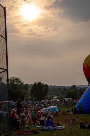 Crowd gathers at sunset hot air balloon festival on green field with colorful balloons inflating under hazy sky.