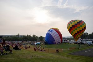 Two colorful hot air balloons inflate at a crowded outdoor festival on a grassy field at sunset.