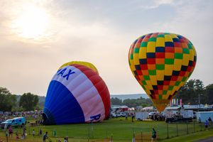 Two colorful hot air balloons, including a RE/MAX balloon, inflate at a festive outdoor balloon festival at sunset.