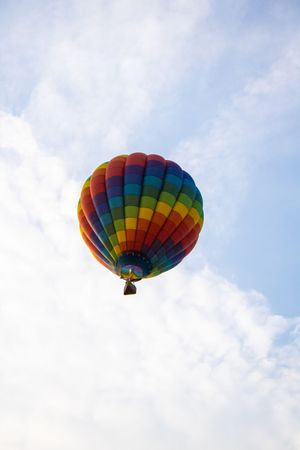 Colorful rainbow striped hot air balloon floating against a partly cloudy blue sky, viewed from below.