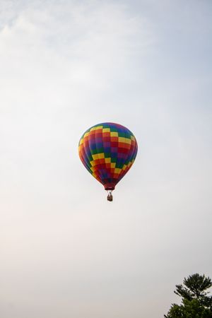 Vibrant multicolored hot air balloon with checkered pattern soaring in hazy sky above green treetops.