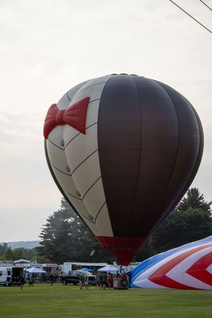 A large dark hot air balloon with a bow tie design inflated at an outdoor festival on a green field.