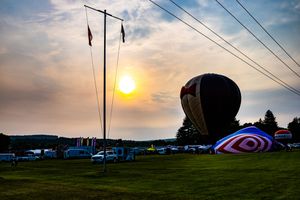 Hot air balloons inflate at sunset during an outdoor festival on a green field with cars and trees in the background.