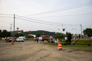 Hot air balloons float over a festive outdoor event with crowds, orange barrels, and green hills in the background.