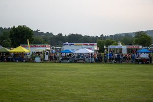 Outdoor food vendor booths lined up on a green field at dusk during a community festival event.