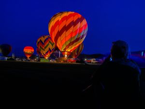 Colorful hot air balloons glowing at dusk during a night balloon festival with spectator silhouette.
