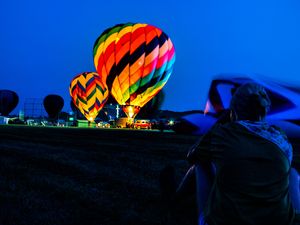 Person photographing colorful glowing hot air balloons at dusk during a night balloon festival.