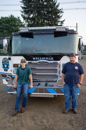 Two people stand proudly in front of a Wellsville fire truck outdoors at dusk.