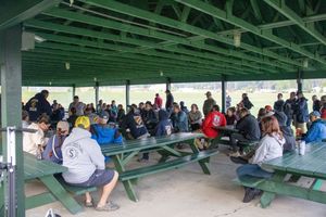 Large group seated at green picnic tables under a pavilion at an outdoor sports venue.