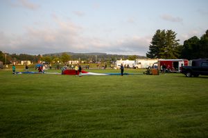 People gather on a lush green field at dusk with vehicles and mountains in the background.