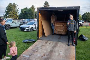 Men unloading a wooden hot tub from a trailer at an outdoor event, with a child and woman watching nearby.