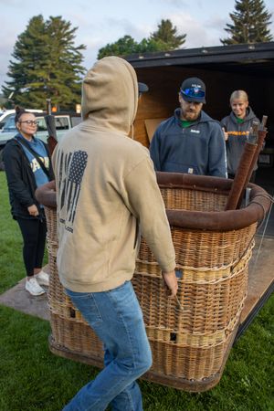People unloading a wicker hot air balloon basket from a truck trailer outdoors.