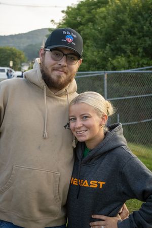 A bearded man in a cap and hoodie poses with a smiling blonde woman in a gray sweatshirt outdoors.