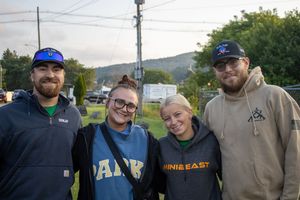 Four friends smile together outdoors at a fall event, wearing casual hoodies and caps with hills in the background.