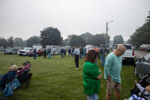 Crowd gathering on a grassy field near parked vehicles and trees on a cloudy day at an outdoor event.