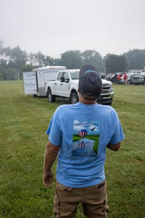 Man in blue Freedom Rises t-shirt stands on grass field at outdoor event with trucks and trailer in background.