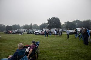 People gather on a grassy field with parked vehicles and trees on a cloudy day at an outdoor event.