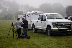 Photographers with tripods set up near a white Ford truck and trailer on a foggy field at an outdoor event.