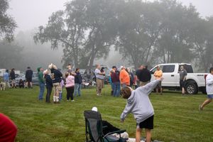 People gather on a foggy morning at an outdoor event on a green lawn with trees in the background.