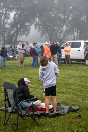 People gathered on a foggy grass field at an outdoor event, with folding chairs and a white truck visible in the background.