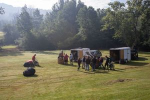 Group of people gathered in a sunny green field near vans and trailers, surrounded by lush trees on a clear day.