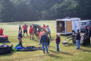 Group of people preparing a hot air balloon in a sunny green field with trailers nearby.