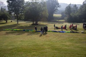 Group of people laying out colorful paragliders on a lush green field with trees and hills behind.
