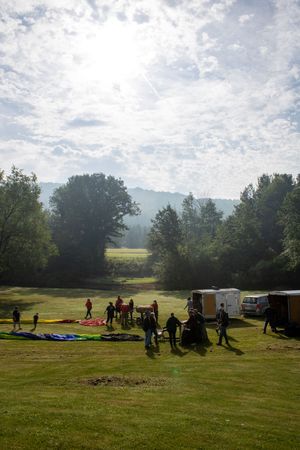 People preparing paragliders on a sunny green field surrounded by trees with mountains in the background.