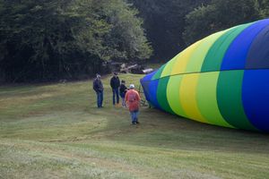 People stand near a large colorful hot air balloon being inflated on a green field surrounded by trees.