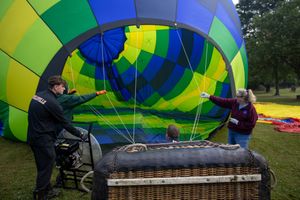Two people inflate a colorful blue and green hot air balloon beside a wicker basket in a grassy park.