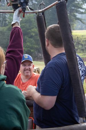 People gathered in a hot air balloon basket, with a smiling man in an orange shirt and blue cap visible.