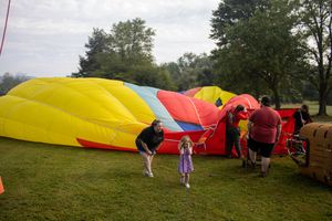 People gather around a colorful deflating hot air balloon in a green park setting on a cloudy day.