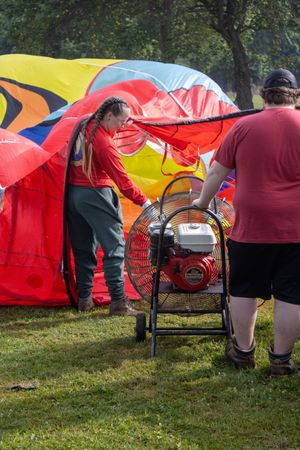 Two crew members inflate a colorful hot air balloon using a large red fan on green grass.