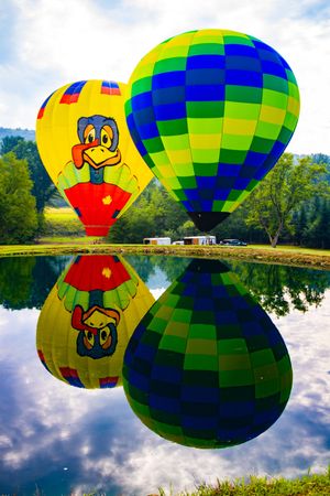Two colorful hot air balloons reflecting on calm water, featuring a cartoon duck design and green checkered pattern.