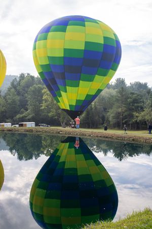 Colorful green, yellow, and blue checkered hot air balloon reflected in calm lake water with trees in background.