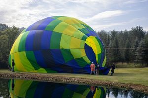 Colorful hot air balloon being inflated on a green field near a reflective pond with trees in the background.