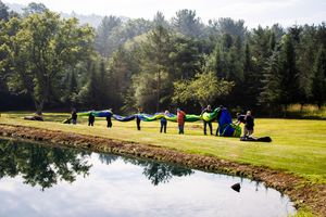 Group of people holding colorful parachute by a reflective pond in a lush green park surrounded by tall trees.
