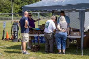 People gathering under a blue canopy tent at an outdoor event on a sunny day, browsing items on a folding table.