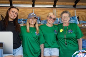 Four women in matching green t-shirts smile together at an outdoor event booth.