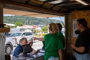 People gathered at an outdoor registration booth, with a person in green shirt pointing directions on a sunny day.